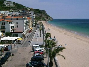 A coastal street with buildings, parked cars, and a beach.