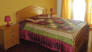 A bedroom with a wooden bed, a nightstand with drawers, a red lamp, and a window with curtains. - Sesimbra Holiday Apartment (Sesimbra)
