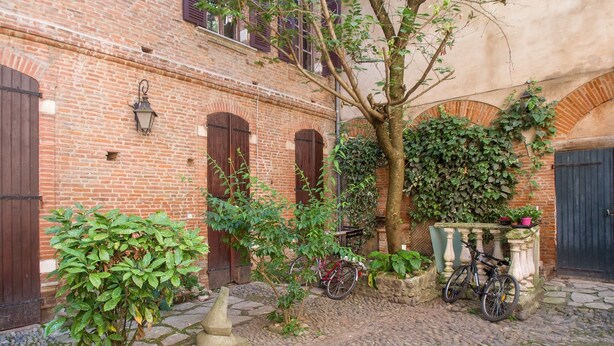 Ein Innenhof mit einem Kopfsteinpflasterweg, einem Backsteinhaus mit Rundbogenöffnungen und einem Baum.