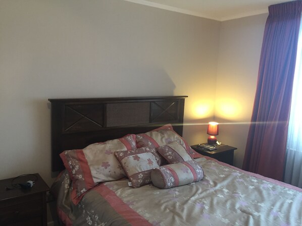 Una cama bien tendida con ropa de cama floral y varios almohadones, una mesita de noche con lámpara y una ventana con cortinas.