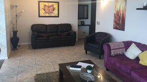 Uma sala de estar com um sofá marrom escuro, um sofá roxo, uma mesa de centro com livros e uma pequena planta, e um quadro na parede.