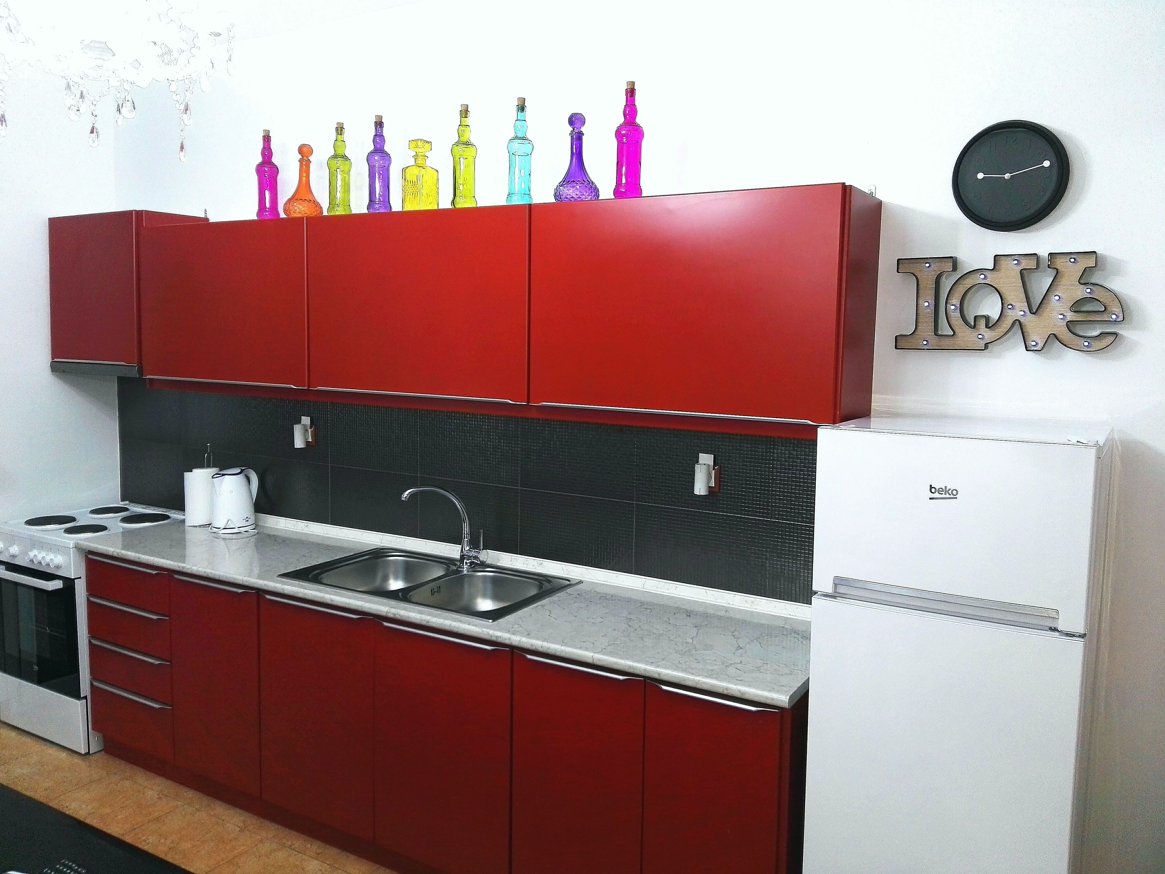 Una cocina con armarios rojos, un refrigerador blanco, un lavamanos y un reloj en la pared.