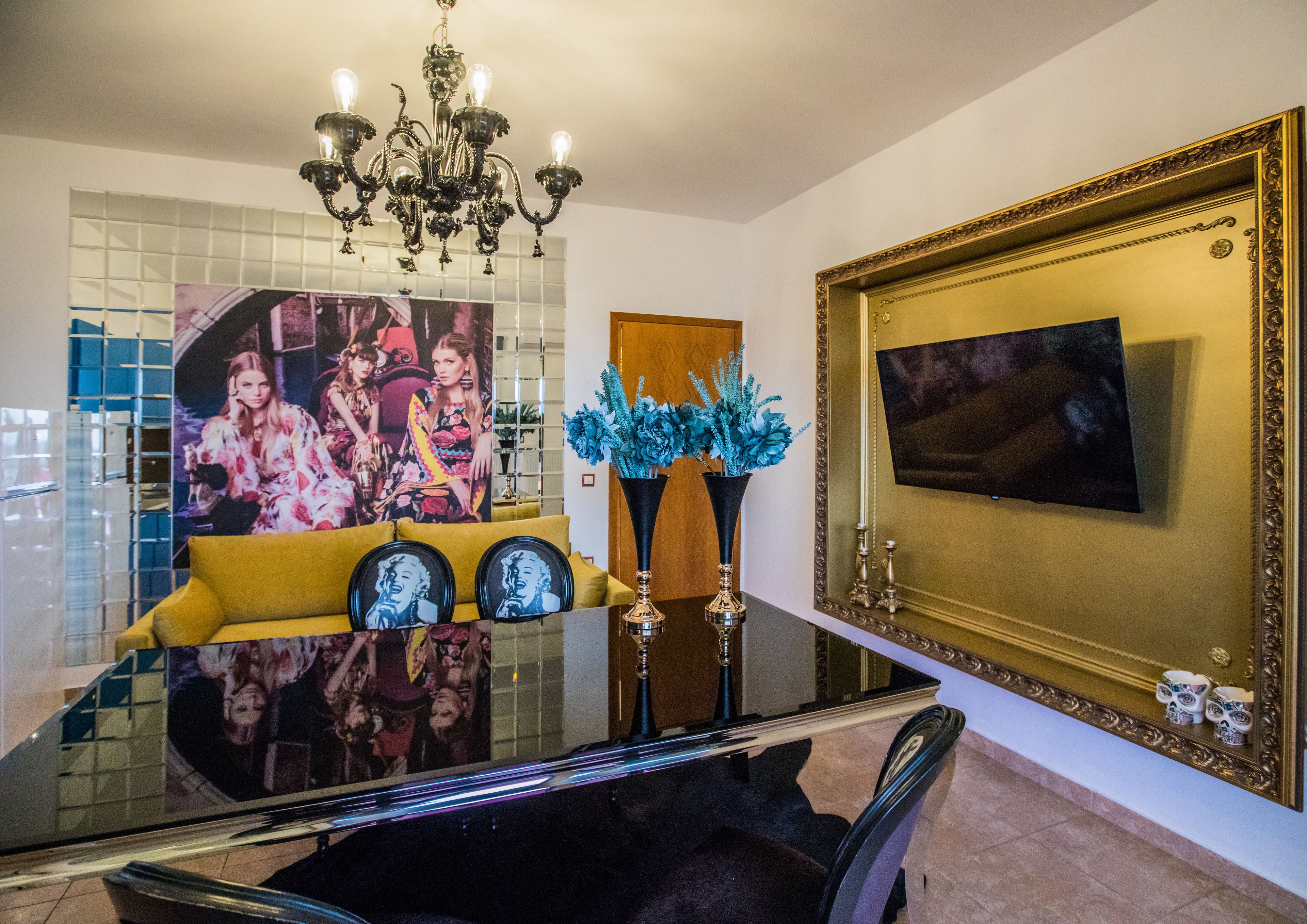 A dining area with a glass table, yellow sofa, blue floral centerpiece, and a large framed mirror.