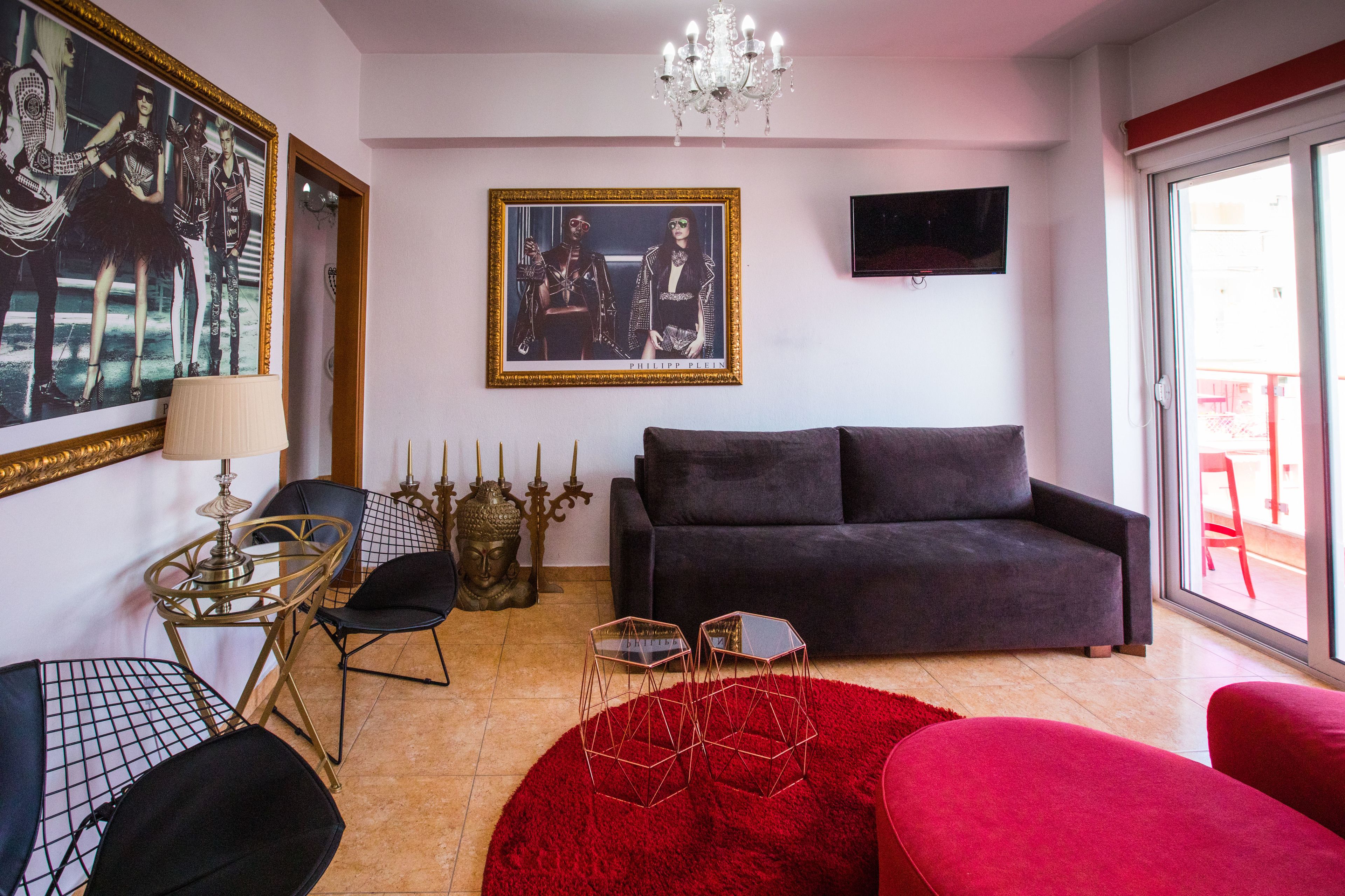 A living room with a dark grey sofa, a red rug, and a chandelier. There are two hexagonal tables with metal frames and a lamp on a small table.