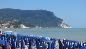 A beach with numerous blue umbrellas and lounge chairs, a clear view of the sea, and a hill with buildings in the background.
