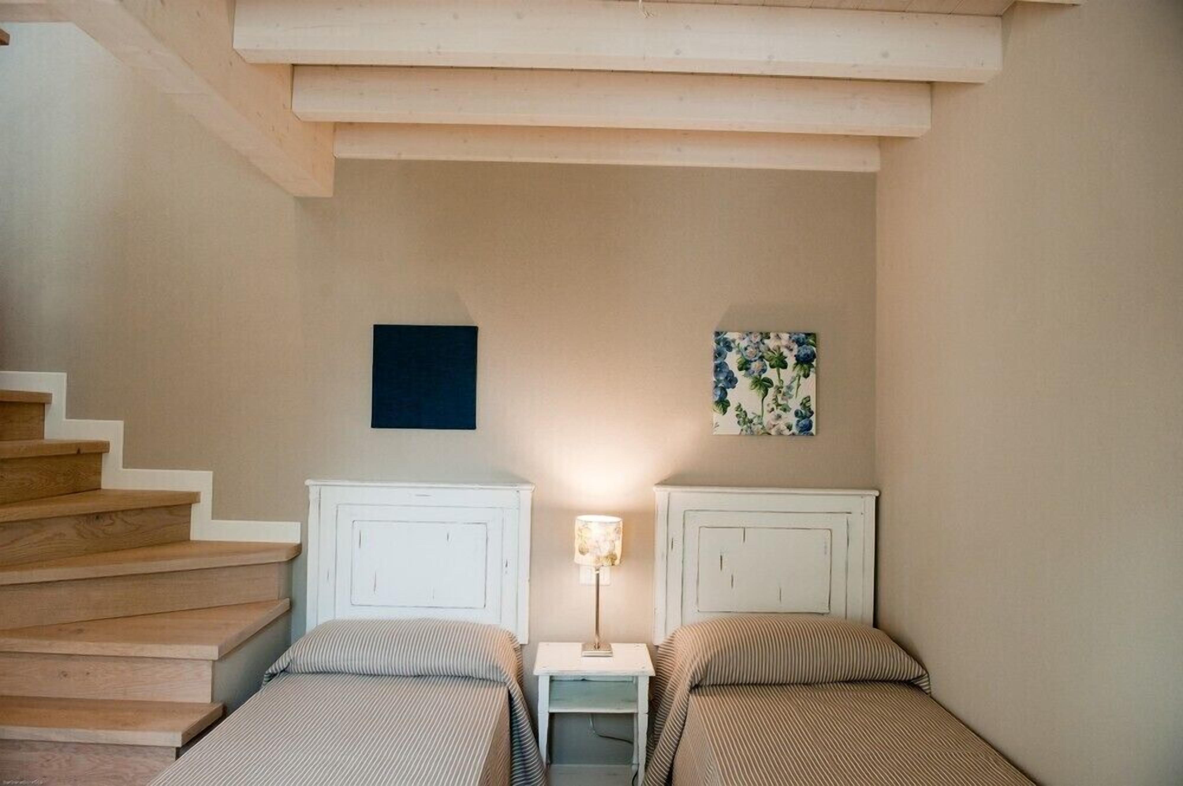 A bedroom with two beds, a nightstand with a lamp, a staircase, and a wall with a framed picture.