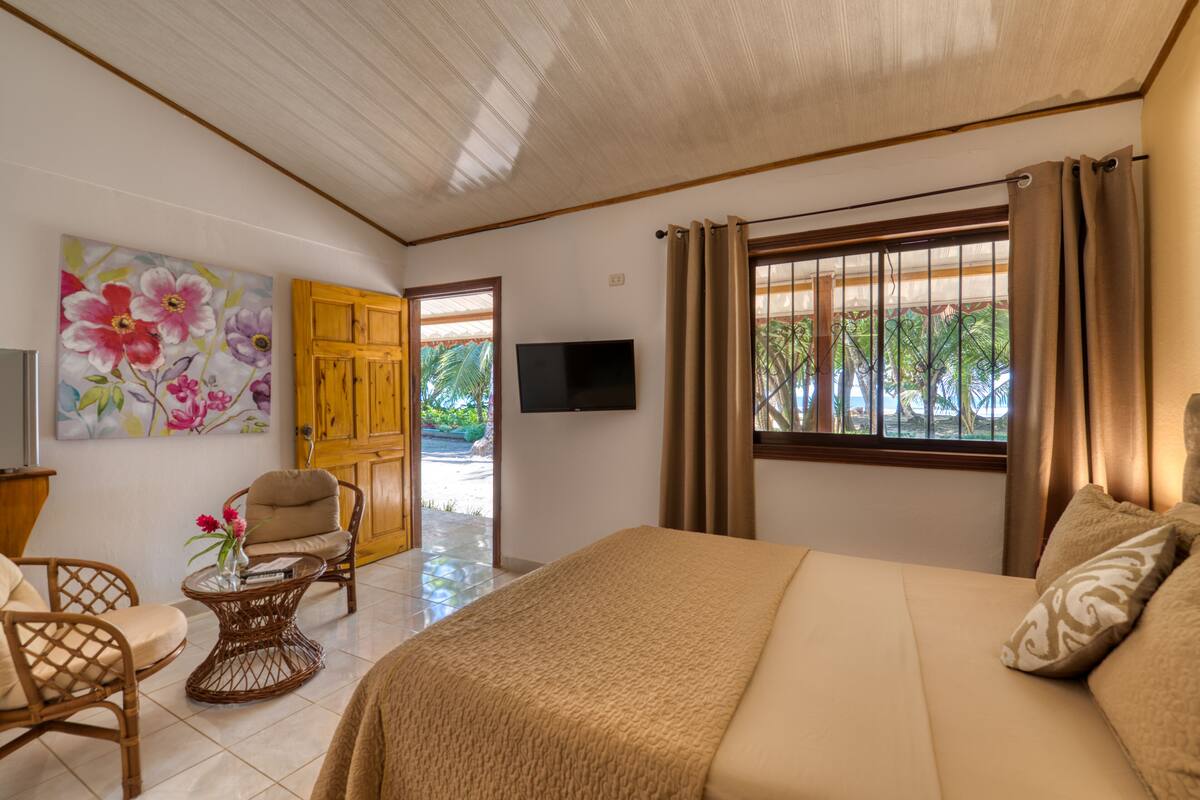 A bedroom with a bed, a television, a chair, a table with flowers, and a door leading outside.