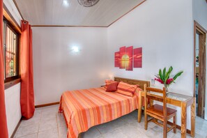 Ein Schlafzimmer mit einem Bett, einem Nachttisch, einem Schreibtisch mit Stuhl, einem Fenster mit Vorhängen und einem Wandbild.