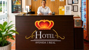A hotel reception desk with a crown logo, clocks displaying different cities, and a reception sign. - Hotel Avenida 3 Real (Cali)