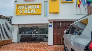 Et hotel kaldet "Avenida 3 Real" med en markant gul og hvid facade, flere vinduer og en bil foran.