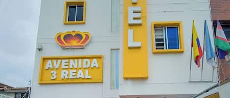 Un hotel llamado "Avenida 3 Real" con una fachada llamativa de color amarillo y blanco, múltiples ventanas, y un automóvil estacionado frente a él.
