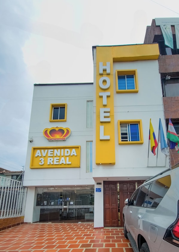 A hotel named "Avenida 3 Real" with a prominent yellow and white facade, multiple windows, and a car parked in front.