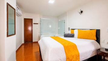Una habitación de hotel con cama, mesita de noche, espejo, puerta y zona de ducha.