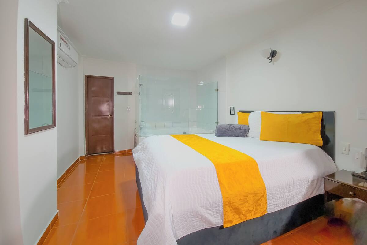 A hotel room with a large bed, a wooden door, a glass shower enclosure, and a white tiled floor.