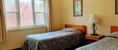 A room with two beds, a window with curtains, a bedside table with a lamp, and a framed picture on the wall.