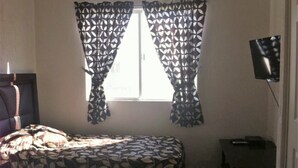 A bedroom with a bed, a window with patterned curtains, a television on the wall, and a small bedside table. - Villas Narcisos (Cancun)
