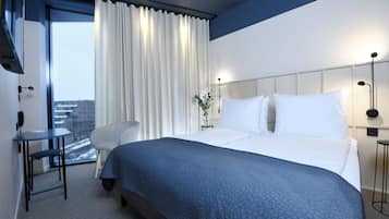 Habitación de hotel moderna con una cama grande, un escritorio pequeño y vistas de la ciudad a través de un ventanal amplio.