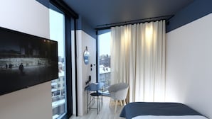 Moderni hotellihuone, jossa on suuri ikkuna, sänky, tuoli, pieni pöytä ja seinälle asennettu televisio.