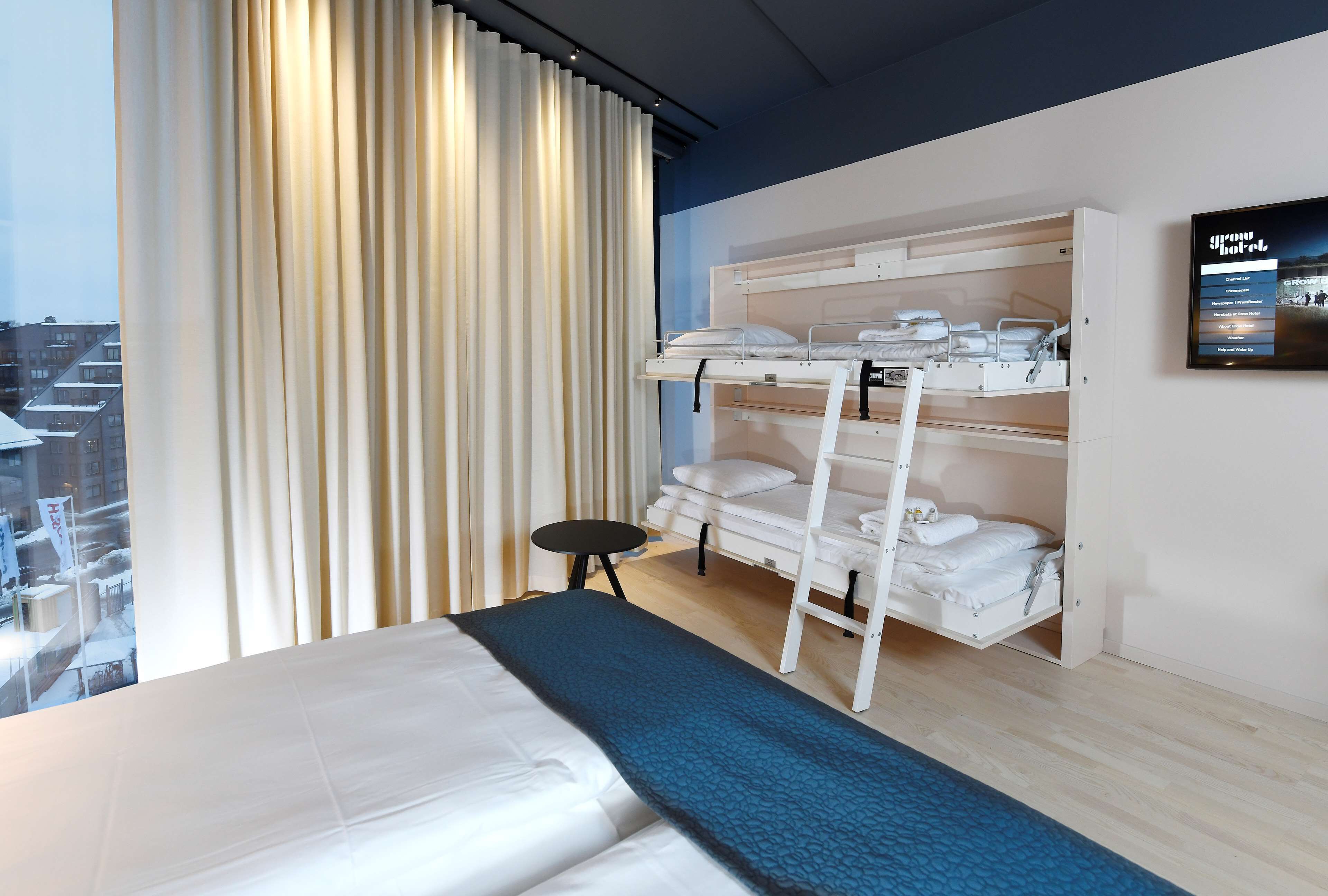 A hotel room with a large bed, a bunk bed with storage, a small table, and a view of the city through the window.