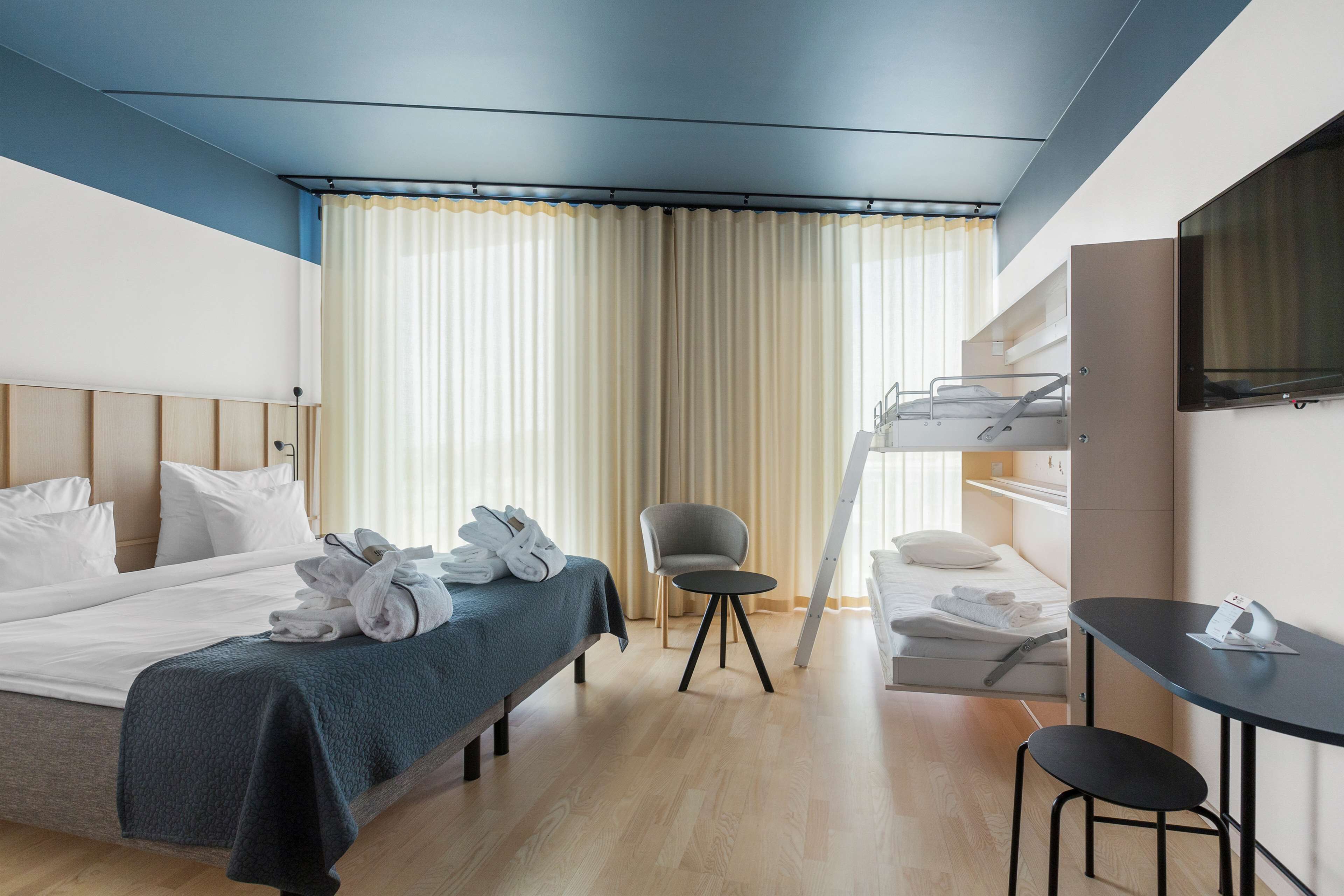 Habitación de hotel con cama, escritorio, silla, televisor y ventana con cortinas.