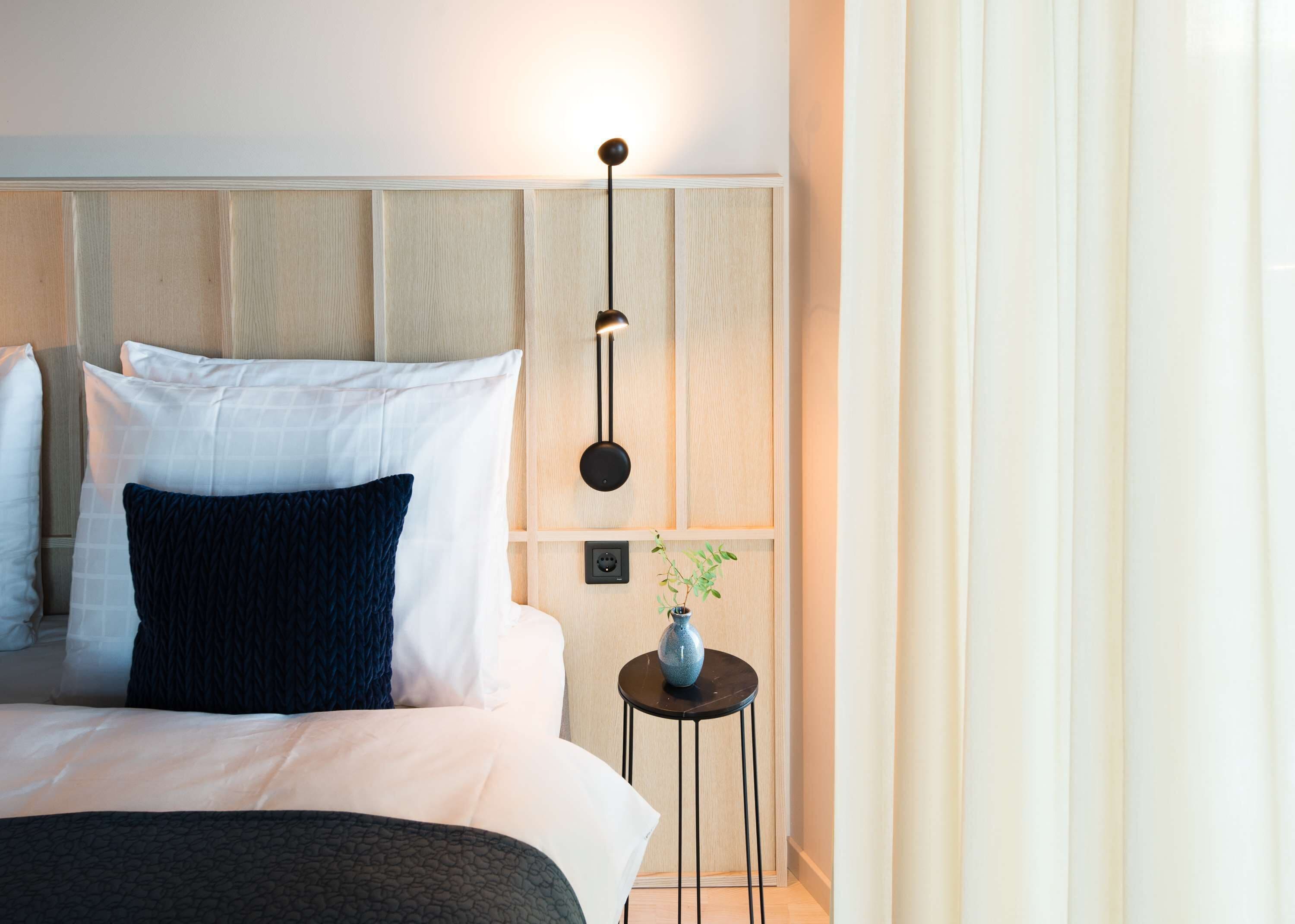 A modern hotel room with a bed, a bedside table with a vase, and a wall-mounted light fixture.