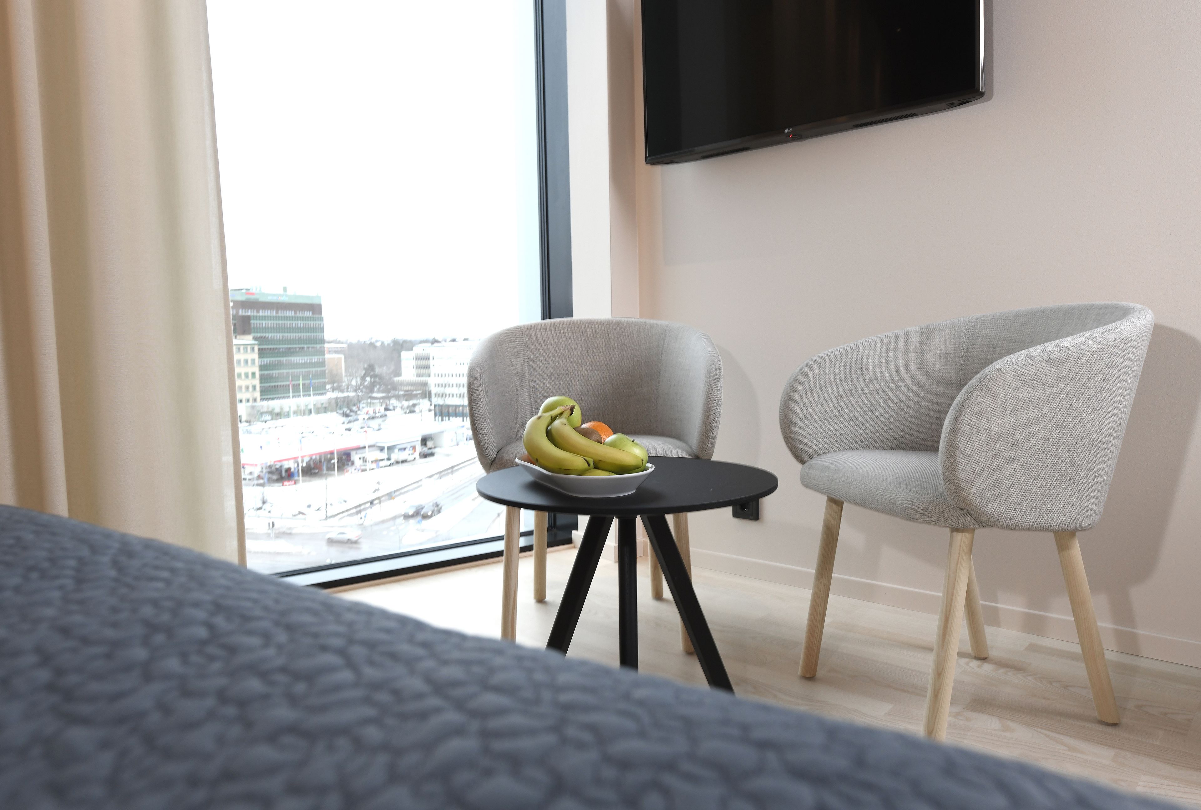 Een hotelkamer met een bed, een klein tafeltje met een schaal fruit, twee grijze stoelen, een aan de muur bevestigde televisie en uitzicht op een stadsgezicht door een groot raam.