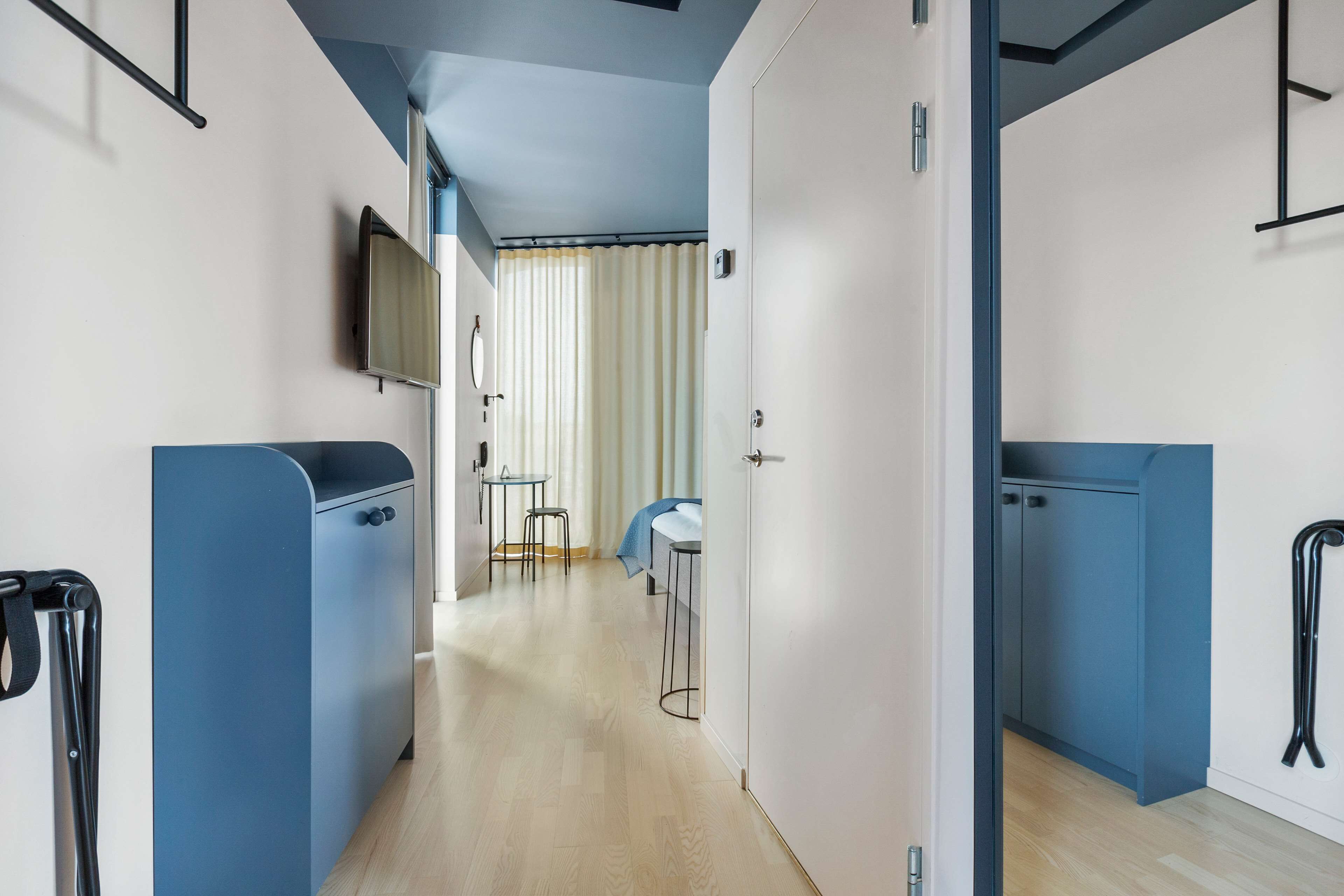 A modern hotel room with a wooden floor, a blue wardrobe, a small table with two chairs, a TV mounted on the wall, and a curtain-covered window.