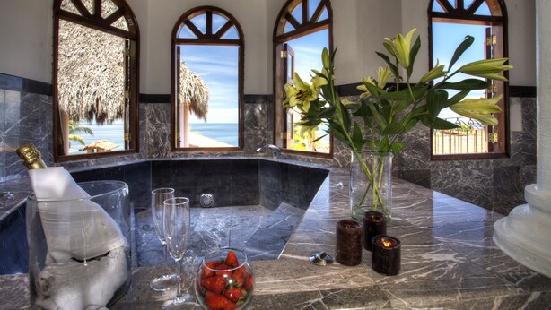 Um banheiro luxuoso com bancada de mármore, uma banheira grande e vista para o oceano através de janelas em arco.