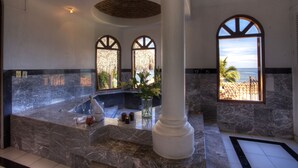 Casa de banho em mármore com banheira, um pilar grande e vista para a praia através de uma janela.