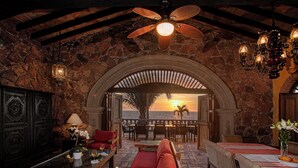 Quarto com paredes de pedra, um sofá vermelho, uma mesa com flores e vista para o pôr do sol sobre o oceano.
