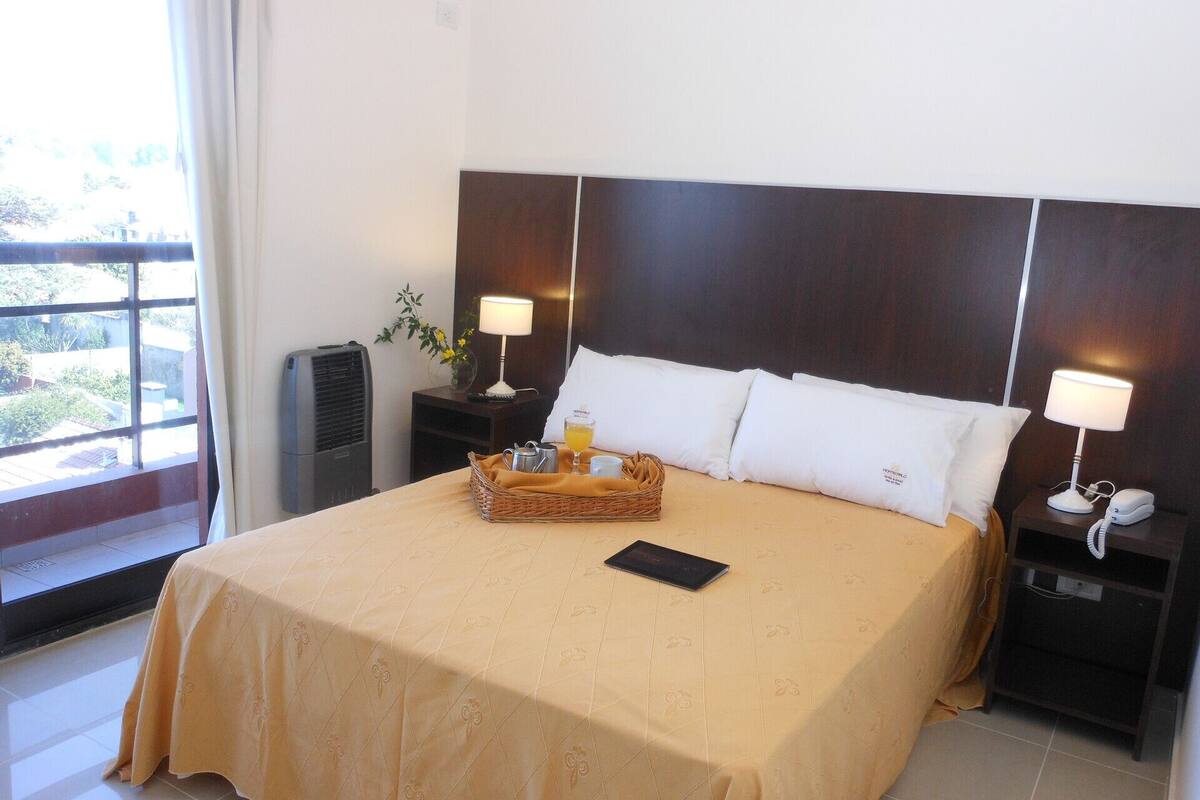 A hotel room with a large bed, two bedside tables with lamps, a small table with a tray of breakfast items, and a window with a view of greenery.