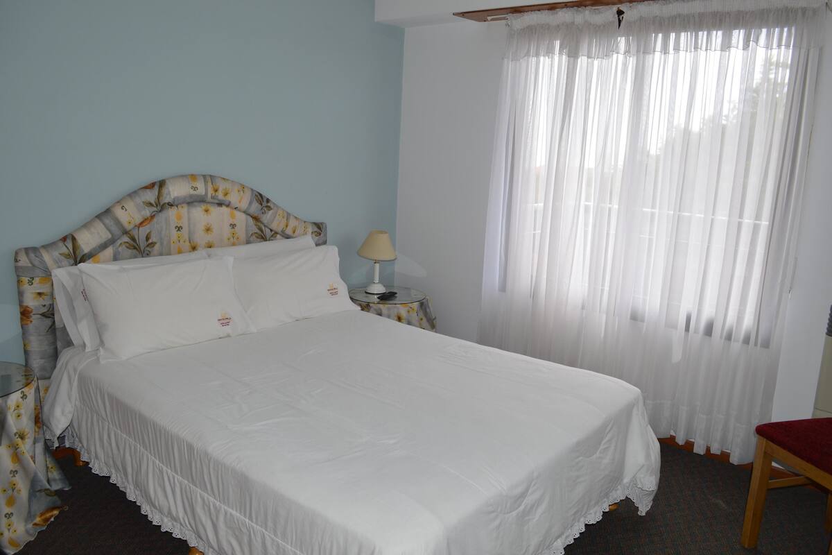 A neatly made bed with a floral-patterned headboard, a bedside table with a lamp, and a chair.