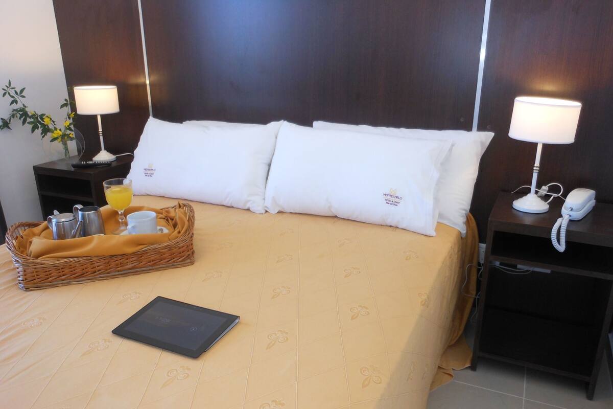 A hotel room with a neatly made bed, two bedside tables with lamps, a bedside phone, a basket with breakfast items, and a tablet on the bed.