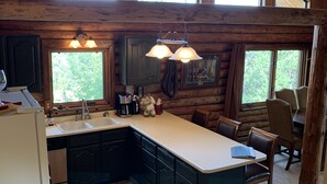 A rustic cabin-style kitchen with wooden beams, a large island, and a view of the outdoors through large windows. - Spa style log cabin retreat on Sandia Mountain (Cedar Crest)