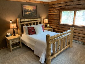 A wooden log cabin bedroom with a bed, two bedside tables, lamps, and a painting on the wall. - Spa style log cabin retreat on Sandia Mountain (Cedar Crest)