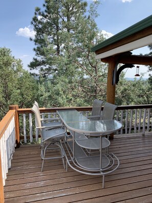 A wooden deck with a glass table and metal chairs, surrounded by trees and a clear sky. - Spa style log cabin retreat on Sandia Mountain (Cedar Crest)