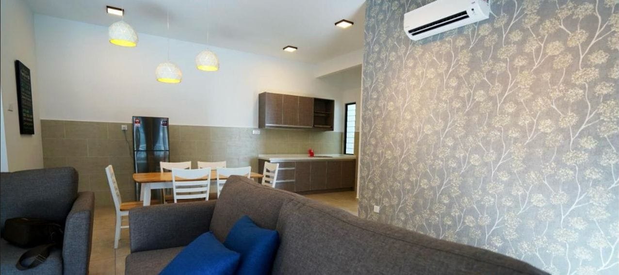 A modern living room with a grey sofa, blue cushions, and a dining area with a wooden table and chairs. There is a kitchen in the background with wooden cabinets and a refrigerator.