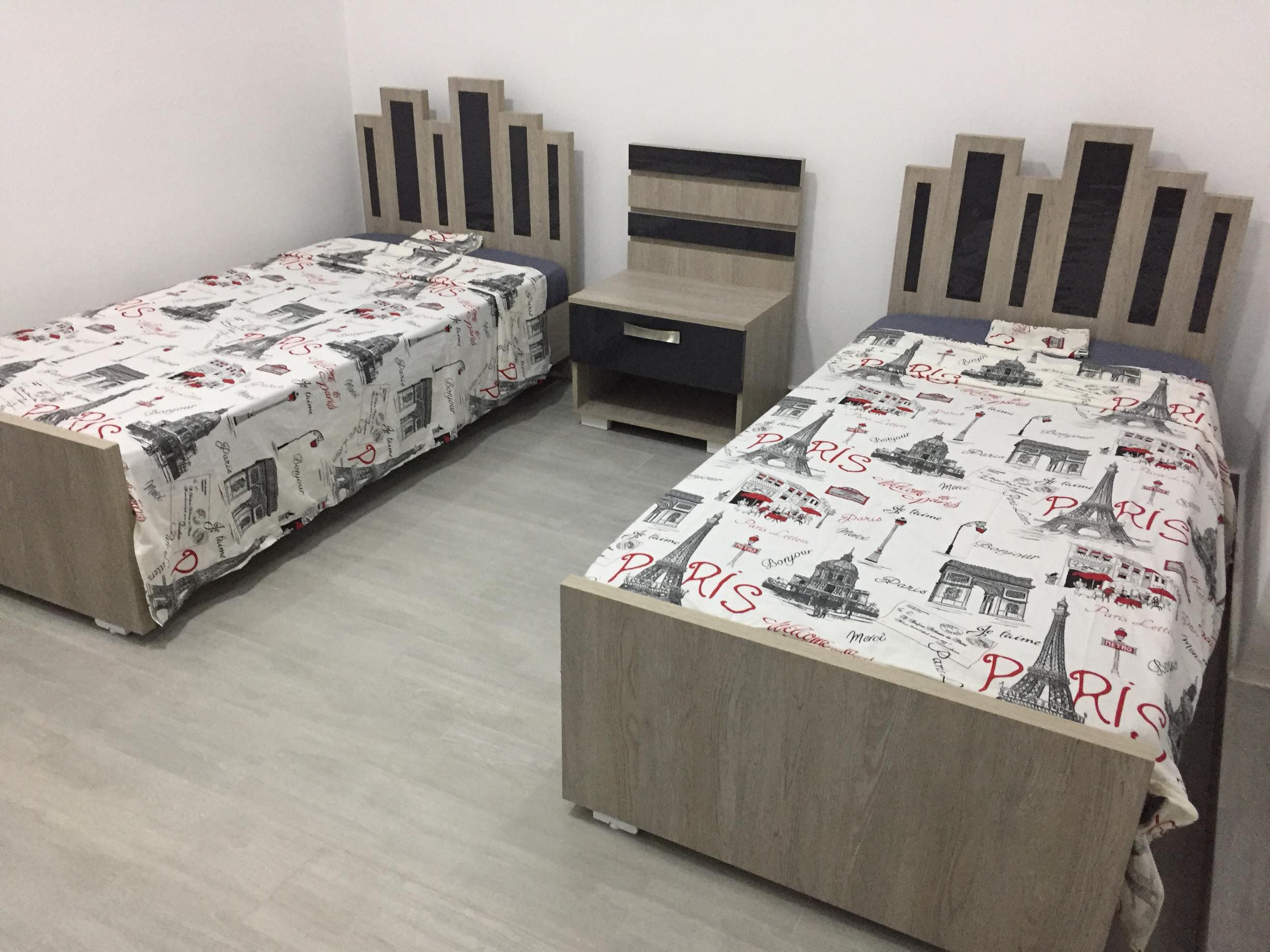 Una habitación con dos camas individuales, cada una con colcha con motivos parisinos. Hay una mesita de noche pequeña con un cajón entre las camas.