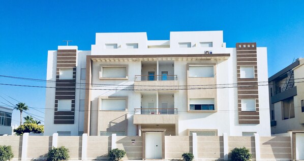 A modern multi-story residential building with balconies, a flat roof, and a gated entrance.