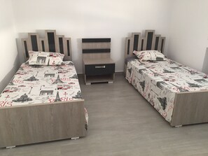 A room with two single beds, each with a Paris-themed bedspread and matching pillows. There is a small bedside table with a lamp between the beds.