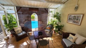 Un patio couvert aménagé avec des meubles en osier, une table et des chaises. On peut apercevoir une piscine à travers une fenêtre.