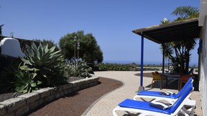 A covered outdoor seating area with two blue lounge chairs and a table, surrounded by plants and a view of the sea. - panoramic view  Vineyards 2 Bed+2bathrrooms Pool Heated  (La Matanza de Acentejo)