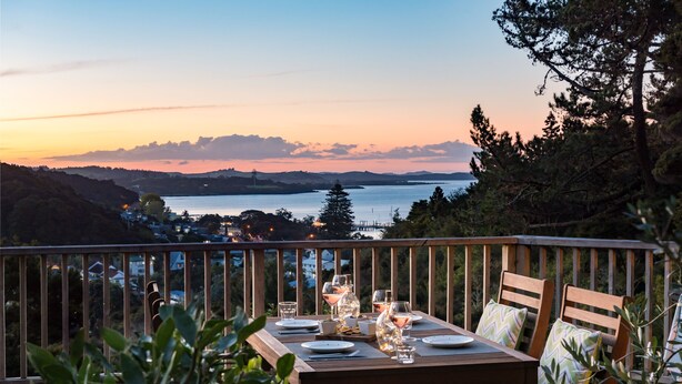 A wooden deck with a table set for dining, overlooking a body of water and distant hills. - Allure Lodge cabin (Paihia)