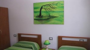 Une chambre avec deux lits, chacun recouvert d’une couverture verte à rayures. Une table de chevet avec une lampe se trouve entre les deux lits. Un tableau représentant un arbre et un paysage est accroché au mur, au-dessus des lits.