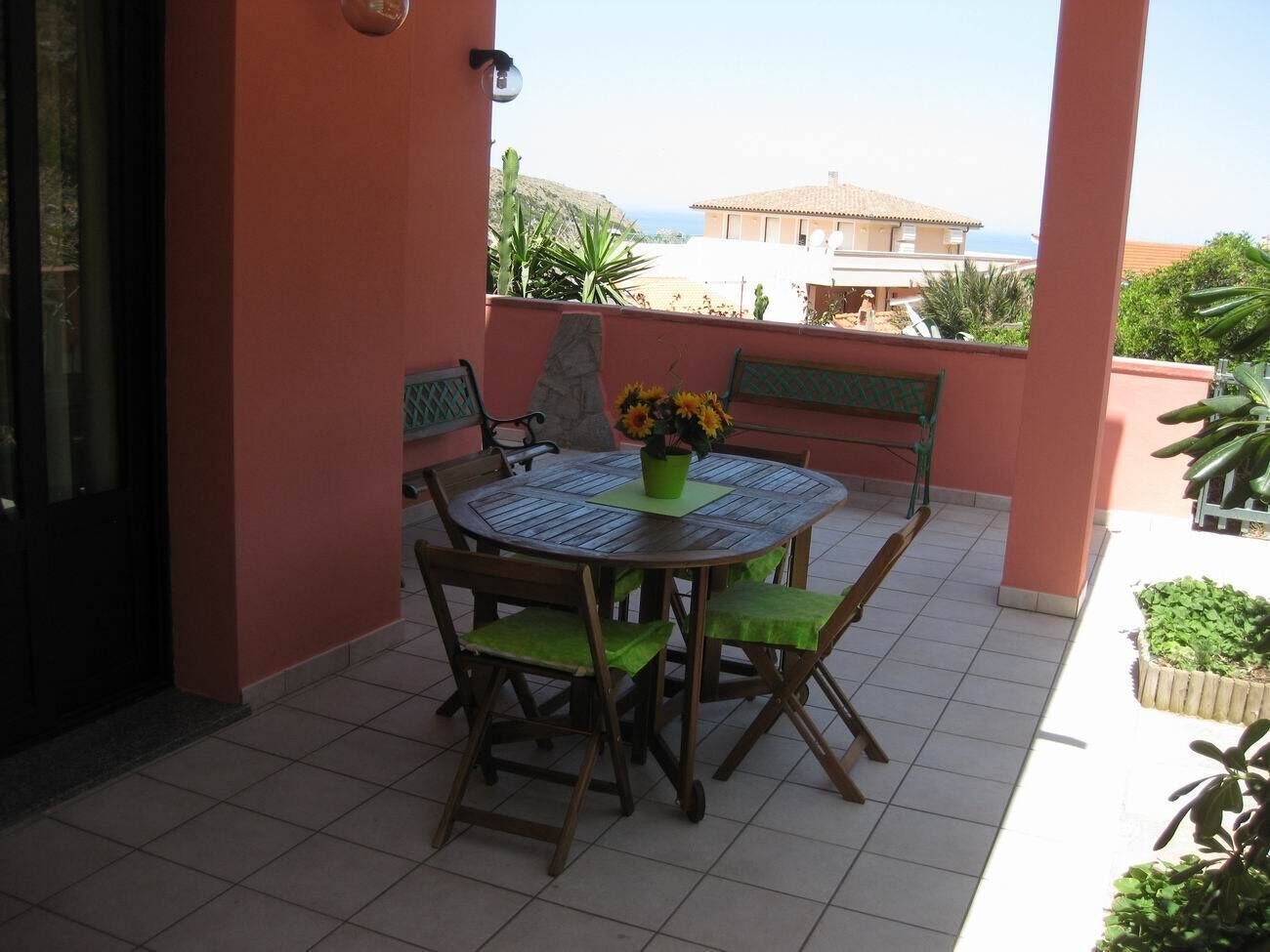 A round table with chairs and a bench, a potted plant, and a view of the sea and buildings in the background.