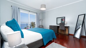 A bedroom with a bed, a desk, a chair, a lamp, a mirror, and a view of the sea. - Casa Eleanor by LovelyStay (Estreito da Calheta)