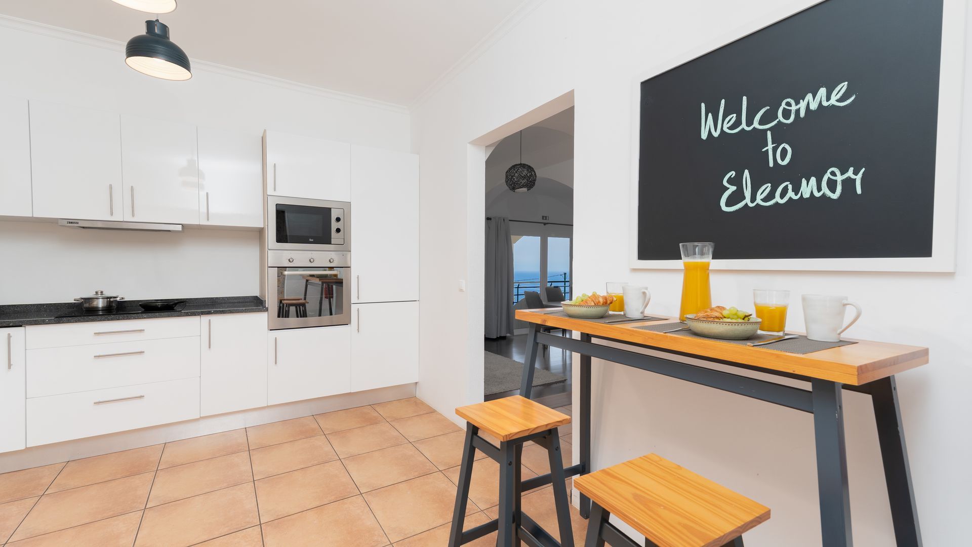 Une cuisine moderne avec des armoires blanches, un four encastré et un tableau noir sur lequel est écrit : « Bienvenue chez Eleanor ».