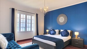 Ein Schlafzimmer mit einem Bett, einem Sessel, einer Chandelüster, einem Nachttisch und einem Wandbild.