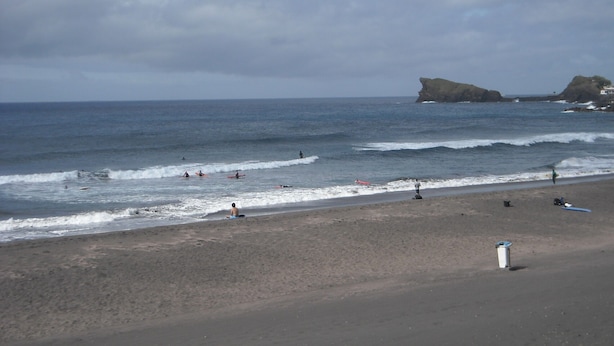 A beach with waves, a rocky outcrop, and a few people.