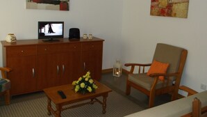 A living room with a television on a wooden cabinet, a floral arrangement on a table, and a chair with an orange cushion.